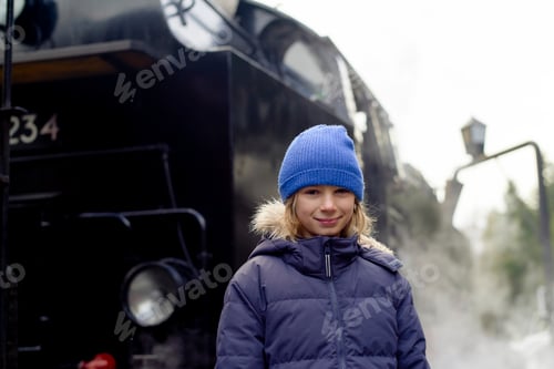 Preview: Child in blue beanie stands smiling by a steaming black locomotive outdoors.