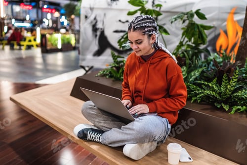 Preview: Young woman using her laptop sitting on bench with her legs crossed in a shopping mall