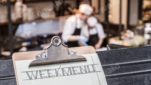 Preview: Week menu clipboard with restaurant kitchen and two cooks blurred busy in the background.