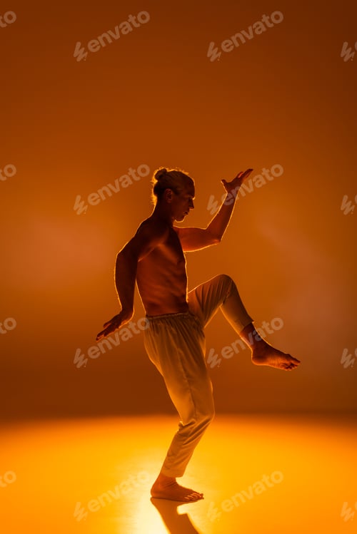 Preview: side view of shirtless man in pants practicing yoga on orange background