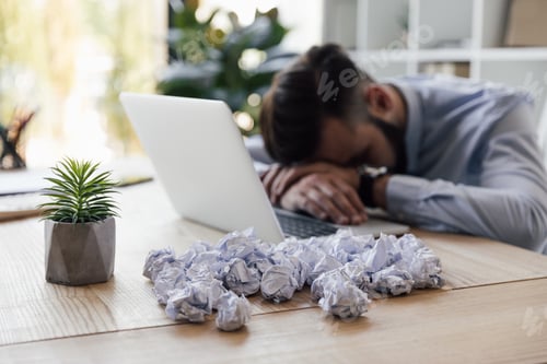 Preview: tired businessman sleeping at workplace with crumpled papers and laptop on it