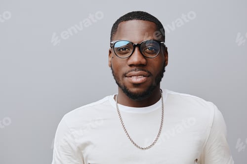 Preview: African man with stylish glasses and a white t shirt, smiling confidently against a soft gray