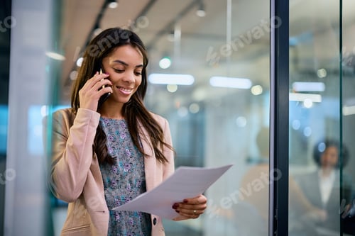 Preview: Happy black businesswoman talking on cell phone while reading documents in the office.