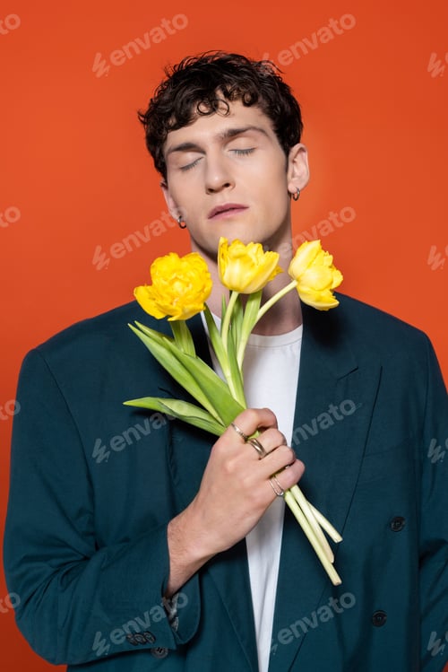 Preview: Brunette man in jacket closing eyes and holding yellow tulips isolated on red