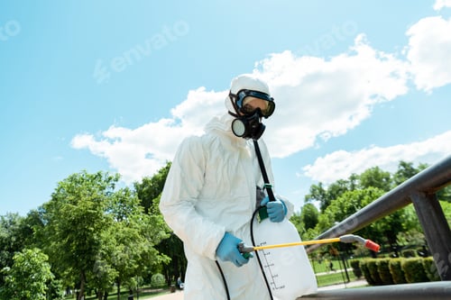 Preview: specialist in hazmat suit and respirator disinfecting railings in park during covid-19 pandemic