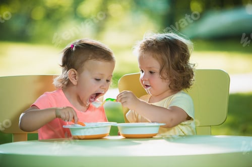 Preview: Two Little 2 Years Old Girls Sitting At A Table And Eating Together Against A Green Lawn