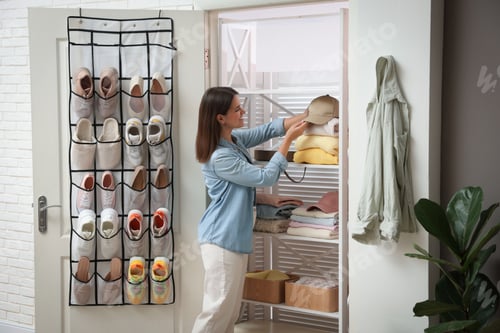Preview: Woman taking cap from shelf near organizer with different footwear at home. Shoe storage