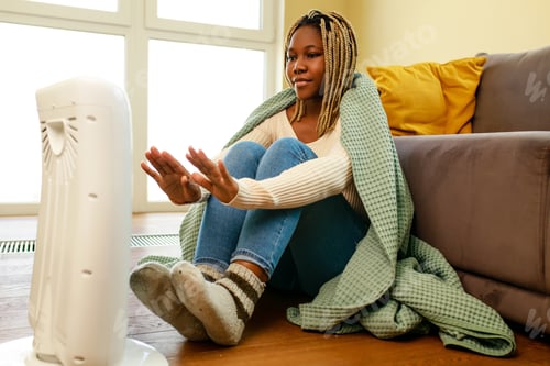 Preview: African american woman in blue blanket warming near electric heater at home