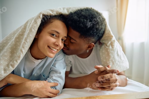 Preview: Young loving couple with bed sheet over their heads in bedroom.