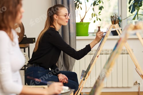 Preview: Young beautiful girl in glasses dressed in black blouse and jeans sits at the easel and paints a