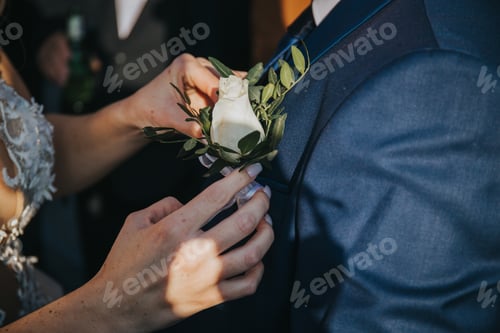 Preview: Closeup of a bride putting a white floral decoration on groom's suit