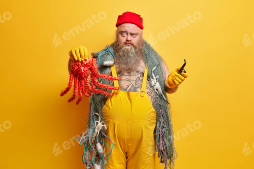 Preview: Strict displeased bearded man seafarer looks seriously at camera holds octopus and smokes pipe goes