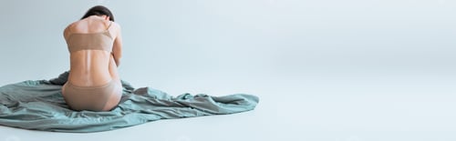 Preview: back view of brunette woman with vitiligo chronical skin condition sitting on blanket on grey