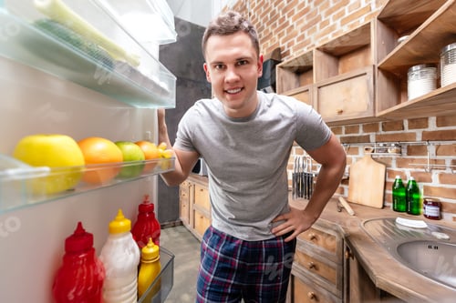 Preview: handsome smiling young man in pajama looking into refrigerator