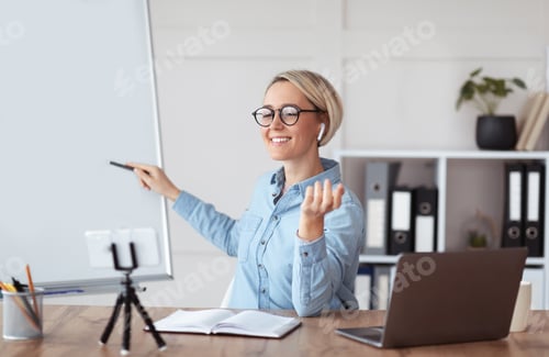 Preview: Positive millennial teacher pointing at empty blackboard near laptop at home, giving online lecture