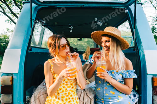 Preview: Two attractive cheerful women drinking lemonade near van, enjoying summer vibes in road trip