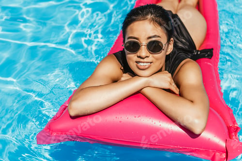 Preview: asian girl in swimsuit and sunglasses sunbathing on inflatable mattress in swimming pool