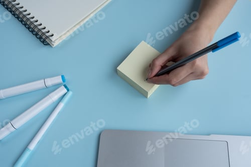 Preview: High angle shot of a person writing on sticky notes and a journal, pens, and laptop on the table