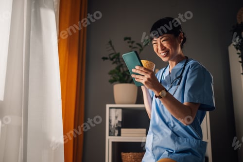 Preview: Asian woman healthcare worker enjoying coffee while checking phone at window