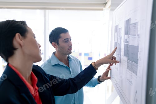 Preview: Side view of Caucasian architects discussing over blueprint on glass board in a modern office