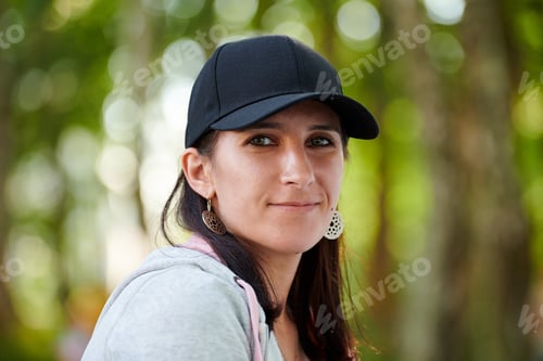 Preview: Young woman with embarrassed smile in black cap and gray hoodie on green forest bokeh background