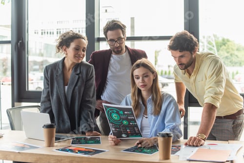 Preview: Businesswoman holding document with charts near interracial colleagues, laptop and coffee in office