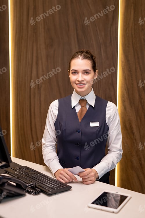 Preview: Happy young brunette female receptionist of modern hotel standing by reception counter