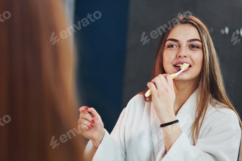 Preview: Beautiful young woman standing in bathroom near the mirror and brushing her teeth