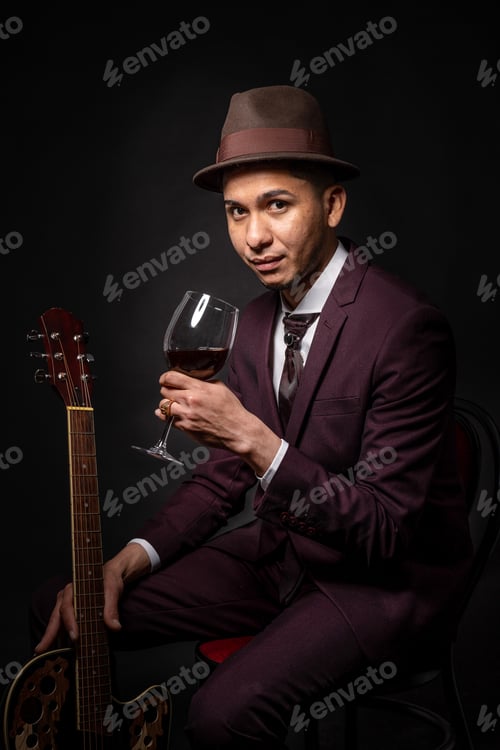 Preview: Latin man in suit with hat and guitar in hand drinking glass of wine sitting and looking at camera