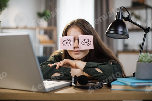 Preview: Tired caucasian female student sleeping with stickers on eyes, sitting at table