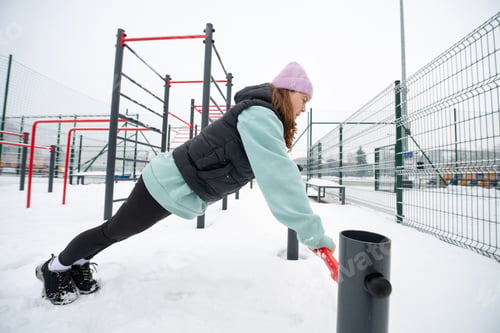 Preview: girl in a push-up position holds onto a horizontal bar on a snowy sports ground
