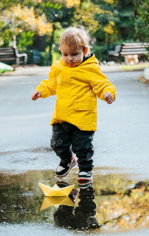 Preview: Cute little boy in yellow raincoat is playing with paper boat in puddle in rainy weather.