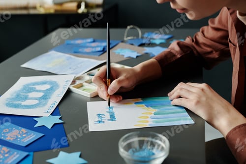 Preview: Young woman with paintbrush bending over desk and painting postcard