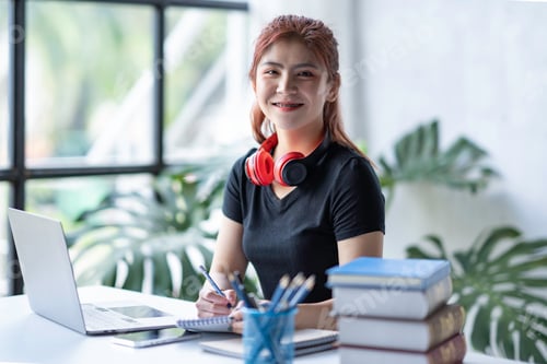 Preview: Asian businesswoman working on a laptop and writing online orders in a notebook in her workstation.