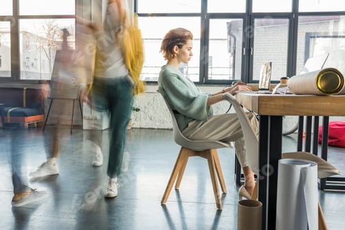 Preview: female architect sitting and using laptop at desk in loft office with colleagues in motion blur on