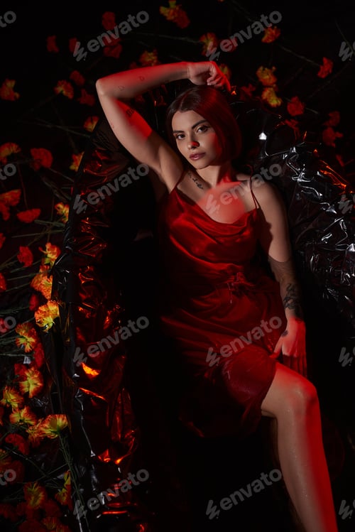 Preview: top view, sensual young woman in red slip dress lying in black bathtub among beautiful flowers
