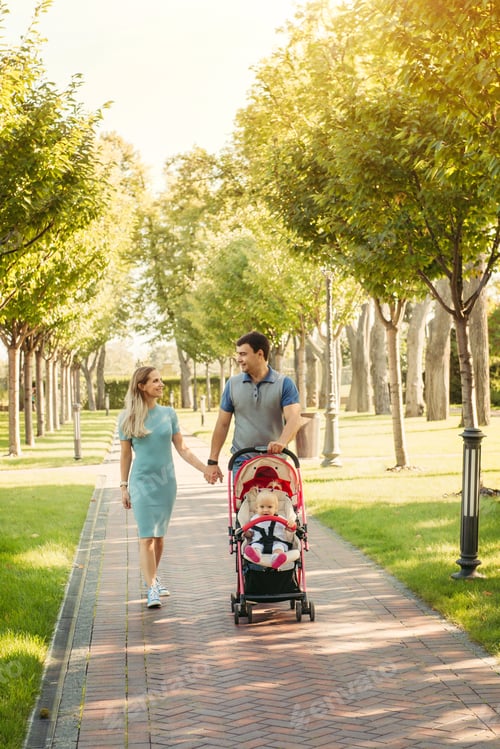 Preview: Young nice couple walking with baby girl in a carriage, looking on each other on green park road