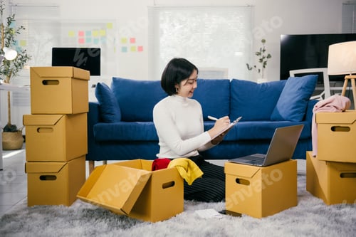 Preview: Asian female entrepreneur checking inventory using laptop and clipboard at home