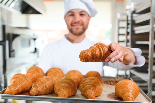 Preview: Handsome baker in uniform holding tray full of freshly baked croissants at the manufacturing