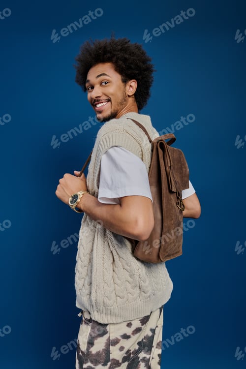 Preview: Young man smiles joyfully while displaying a stylish backpack against a vibrant blue backdrop
