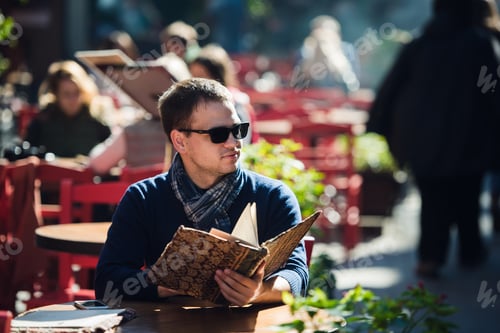 Preview: Young handsome man wearing sunglasses reading the morning paper in a street cafe looking sideways