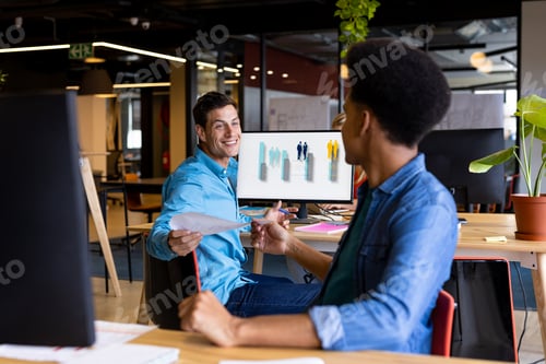 Preview: Happy diverse male colleagues at desks passing document in office