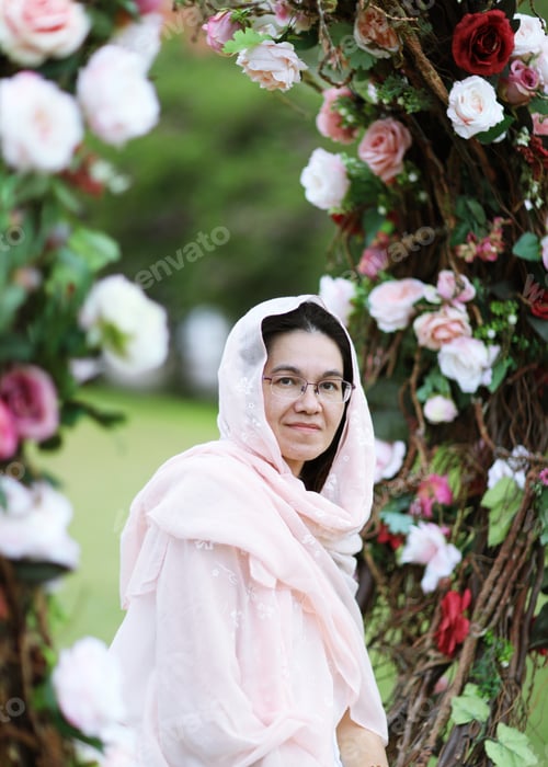 Preview: Woman with pink scarf on head in hot sun sit in the rose arch at the park.