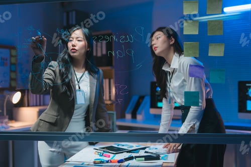 Preview: Businesswomen writing business plan on transparent board in office at night