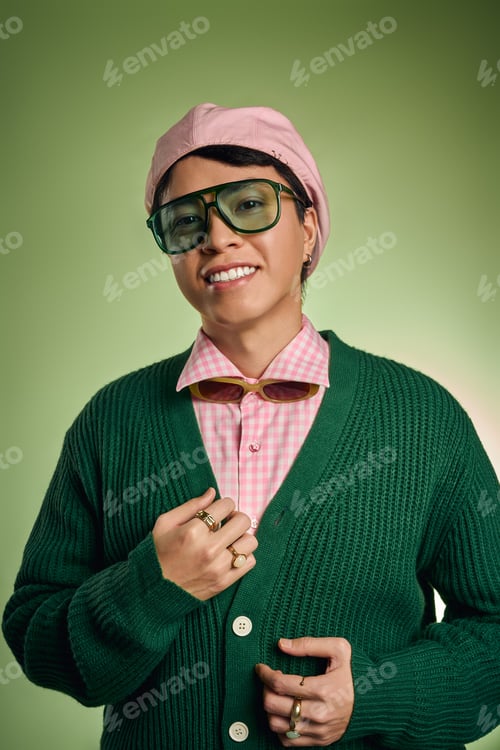 Preview: Stylish man in pink checkered shirt and cardigan poses in vibrant green studio