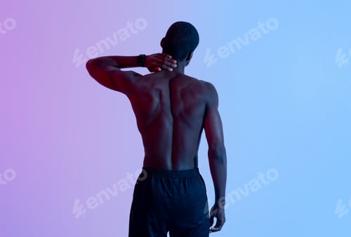 Preview: Back view of young black sportsman with naked torso rubbing his neck, suffering from pain in neon