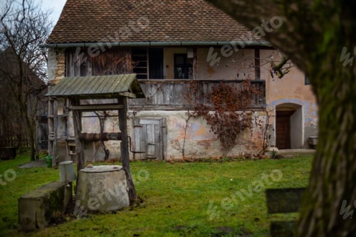 Preview: Scenic view of an old rural house with a green lawn and bare trees on a cloudy day
