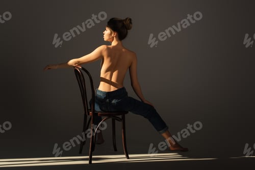 Preview: back view of half nude barefoot woman in jeans posing on wooden chair on grey background with