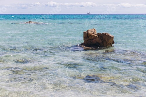 Preview: Costa Rei Sardinia turquoise sea with rocks and distant sailboat under a blue sky