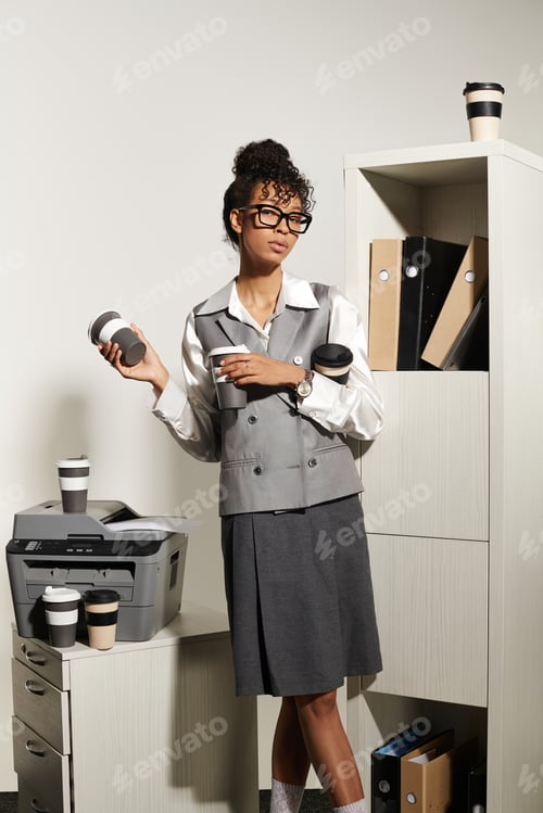 Preview: Elegant young woman in chic business attire, holding coffee cups in a modern office